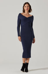 A woman is wearing a form-fitting, long-sleeved knit dress in a deep navy blue. The dress features an off-the-shoulder neckline with a broad, folded collar and has textured ribbing throughout. She completes the outfit with black ankle boots.